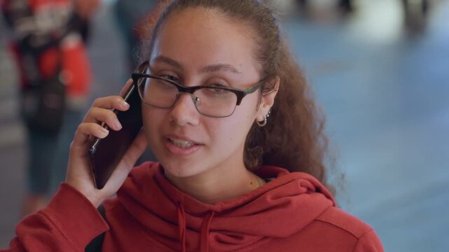 Daytime street phone call young woman in red hoodie animated expressions while navigating crowded plaza candid closeups show decisive gestures and focused speech, glasses, ponytail, blurred scooters