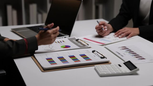Businesswoman discussing financial charts during a late night meeting. Two dedicated female colleagues working overtime in office, analyzing financial data on documents and a laptop.