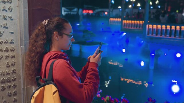 Young woman filming blue fountain show stone railing, yellow backpack, red hoodie, handheld smartphone recording light reflections on water, crowd silhouettes, joyful expression, travel documentation,
