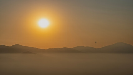 Obraz premium Mountain sunset. The sun is low, shining in an orange sky. There is a silhouette of a flying balloon above the ridge. Evening haze, fog in the valley. Nepal. Sarangkot. The Himalayas.