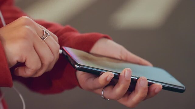 Closeup hands holding smartphone earphones red hoodie, tapping touchscreen to navigate playlist, visible charging cable and silver rings, soft bokeh city lights, evening commute vibe, casual manicure