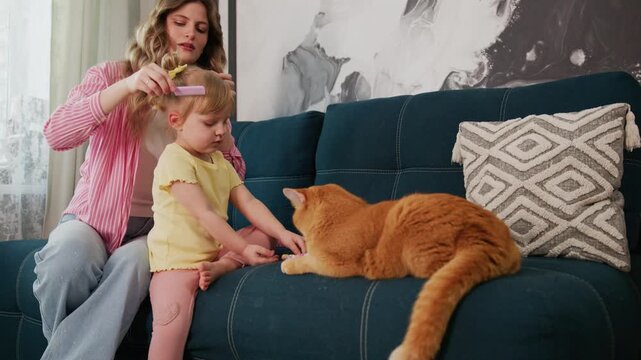 Mother gently brushes her young daughter's hair while seated on a blue sofa. The child smiles, holding a small toy, enjoying the moment. Large orange cat lounges nearby, eyes half-closed, relaxed