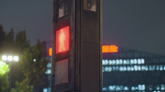 Red pedestrian signal at night glowing on metal pole beside illuminated office building and trees, soft bokeh lights and distant signage create moody urban caution, clear stop message for pedestrians