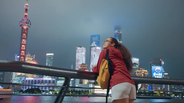 Young black woman leaning on railing overlooking shanghai riverfront with neon towers and Oriental Pearl Tower visible through mist, red jacket and yellow backpack, contemplative backpacker absorbing