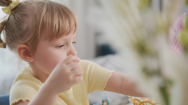 Young girl with blonde hair in a ponytail eats happily. She wears a yellow shirt, focused on her snack. Soft lighting and blurred flowers create a cozy, warm atmosphere
