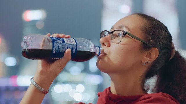 Young woman with glasses, city skyline at night, traveler pauses on waterfront to sip bottle and absorb neonlit skyline, bustling bokeh background, red hoodie, backpack hint, candid urban wonder