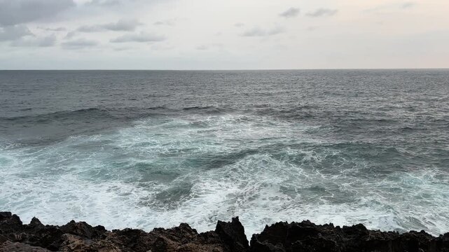 Dynamic seascape showing turquoise waves crashing against dark volcanic rocks with vast gray ocean stretching to horizon under overcast sky, creating dramatic coastal scene.