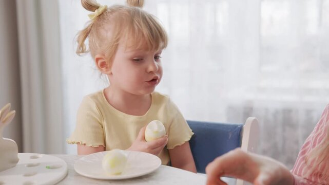 Young child eats Easter egg with mom nearby. Child focuses on egg, tongue out slightly. Mother smiles, holds another egg. Bright natural light fills room