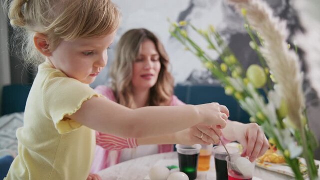 Young girl focused on painting eggs. Mother smiles nearby, guiding her. Bright, colorful dyes on table. Warm, joyful moment of family bonding. Perfect for Easter, crafts, or parenting content