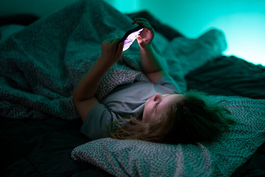 Child holding smartphone under blanket light. Kid using technology during bedtime routine. Child watching video on phone at night. Kid scrolling content on mobile device in bed.