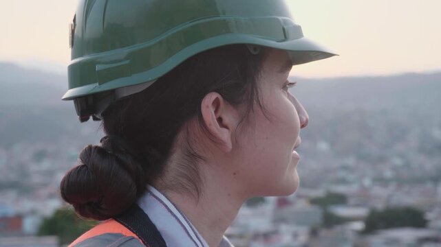 Female construction worker wearing a safety hard hat looking out over the city at sunrise, showing a close-up of her profile while she is thinking and planning her workday