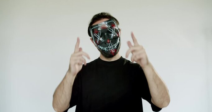 A man wearing a black and red LED purge mask makes shooting gestures with his hands. He is wearing a black t-shirt, and stands in front of a white background. danger, mystery, Halloween, or dystopia.