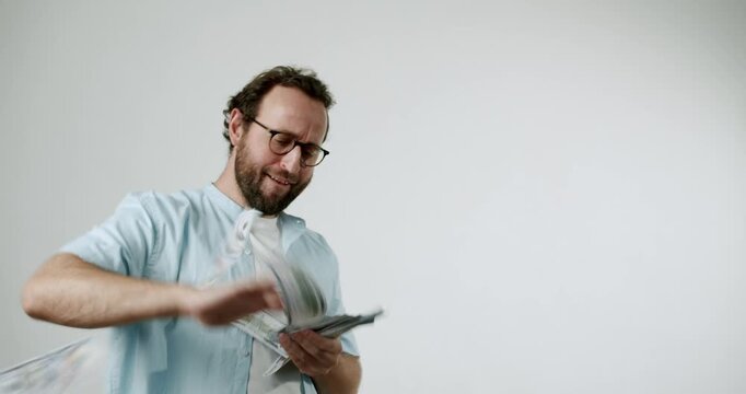 A man throws money in the air. He is wearing glasses and a light blue shirt. He has a beard and is making a funny face. The background is white. This video evokes feelings of wealth and success.