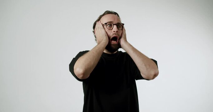 A surprised man in a black t-shirt and glasses reacts to unexpected news. He holds his head in his hands and opens his mouth in shock. His expression conveys disbelief and astonishment.