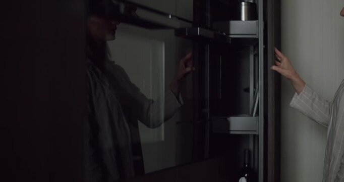 A person is seen opening a sleek, modern glass cabinet in a dimly lit room, creating a reflective and mysterious atmosphere. Person Opening a Modern Glass Cabinet in a Dimly Lit Room