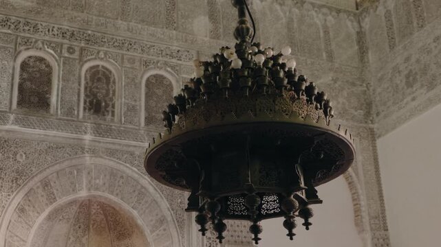 Ornate Chandelier Hangs From Ceiling of Building in Fez, Morocco. Close-up of an old, ornately carved chandelier inside a Moroccan building. The light fixture with small white round bulbs hangs down