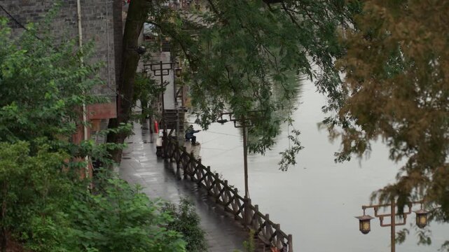 Riverside walkway in rain, asian commuter, slow stroll along wet stone path lined with wooden railing and vintage lanterns, overcast sky and mist, lush greenery and brick wall, distant red coat,