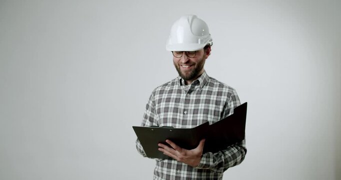A smiling engineer in a white hard hat and glasses writes in a folder on a white background. He makes notes, plans, and takes notes. He is satisfied with his work. Engineering, construction,