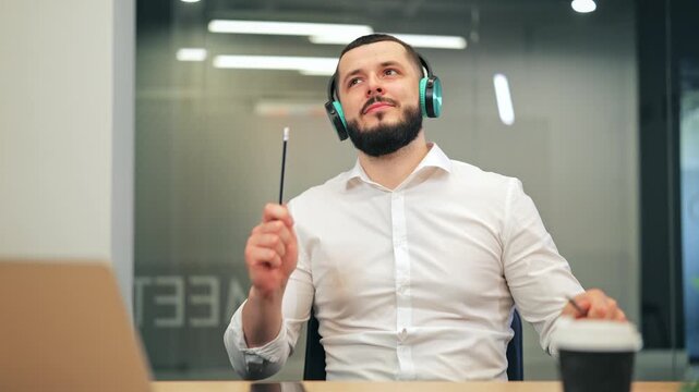 Bearded worker in white shirt enjoys music with headphones, conducting imaginary orchestra using pencils. Happy and relaxed, taking a creative break in the office environment, feeling inspired.