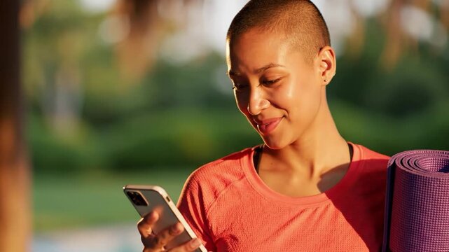 Smiling woman with buzz cut holding phone and yoga mat outdoors