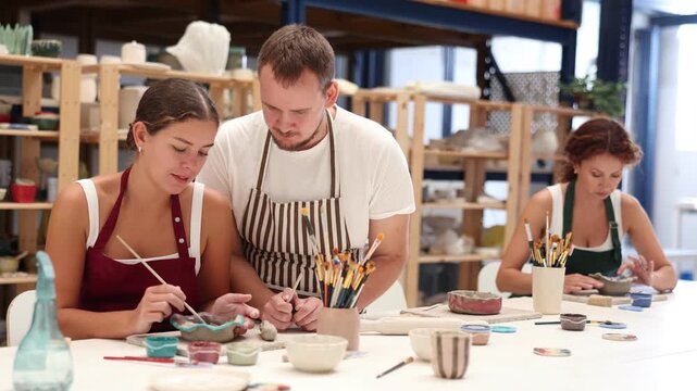 Experienced potter helps students learn how to make patterns on ceramic plates using paint and brush in a pottery workshop. High quality 4k footage