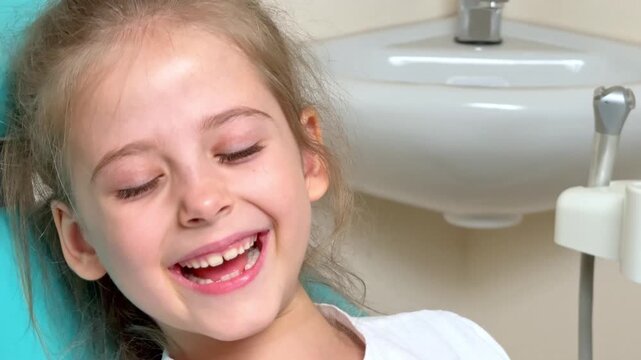 Happy laughing girl with light brown hair sitting in a dental chair inside a modern dental clinic with dental tools visible in the background