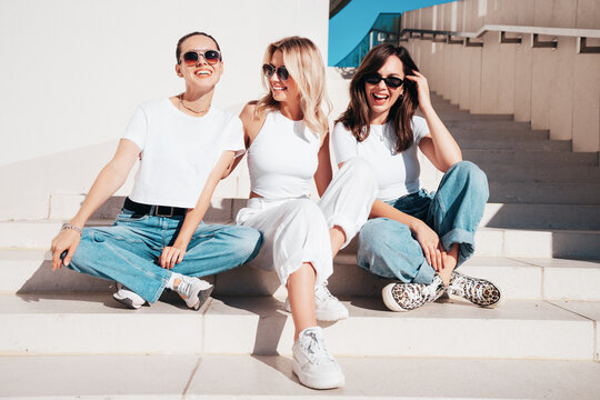 Three young beautiful smiling hipster female in trendy summer t-shirt  and jeans clothes. Sexy carefree women posing in street. Positive models having fun, sit at stairs. In sunny day, sunglasses