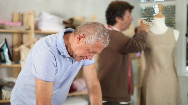 Mature tailor draws a sketch and pattern of clothes on a brown paper next to the assistant. Process of creating clothes in a sewing workshop. High quality 4k footage