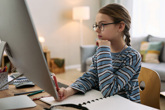 Caucasian girl child wearing glasses sitting at desk using desktop computer while writing in notebook, focusing on screen, studying or doing homework in home environment