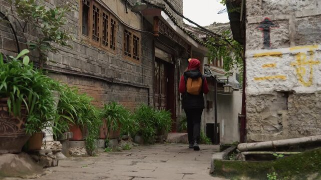 Backpack clad woman walking narrow stone alley toward distant doorway, potted greenery lining brick wall, steady pace and hood up, worn pavement and mossy texture, quiet urban solitude, overcast