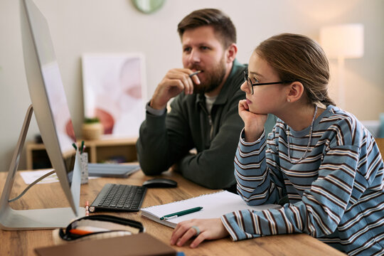 Caucasian middle aged man and Caucasian teenage girl sitting at desk using desktop computer together, both focusing on screen, teenage girl wearing glasses, notebooks and keyboard visible