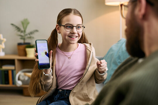 Caucasian girl child with long hair wearing glasses smiling and showing smartphone screen to bearded Caucasian man sitting nearby, demonstrating new mobile application