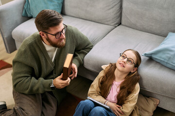 Caucasian middle aged man sitting on floor holding book talking to Caucasian preteen girl, lying on carpet smiling and looking up wearing glasses in living room setting