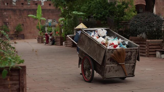 Waste collector pushing loaded cart through brick courtyard filled with plastic bags and greenery, wearing conical hat, rustic metal cart with rusty wheel, vendor stalls in background, quiet morning