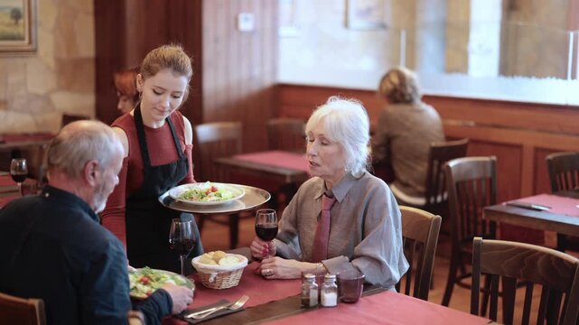 Young woman waitress brings order from couple elderly man and elderly woman in restaurant