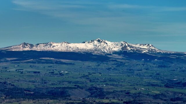 Panoramic view of snow capped Monts Dore mountains in Massif Central France rising above rolling green hills and rural landscape under clear blue sky