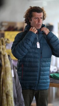Man try on down jacket in front of mirror in clothes store. Customer is trying on clothes in store. Buyer is immersed in choice of clothes. 