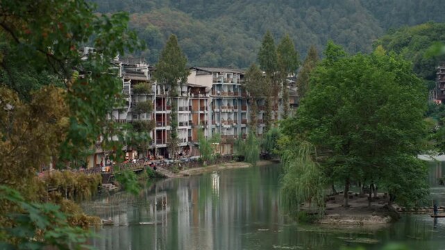 Riverfront buildings with island tree, panoramic waterside town with treetopped islet, broad river reflections, multistorey facades, terrace cafes and moored boats, distant wooded hills and gentle