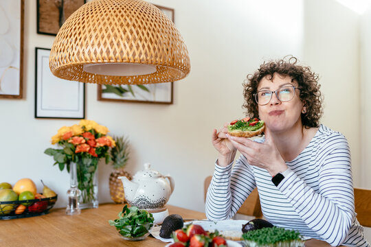 Middle aged woman eating avocado toast smiling at home kitchen table