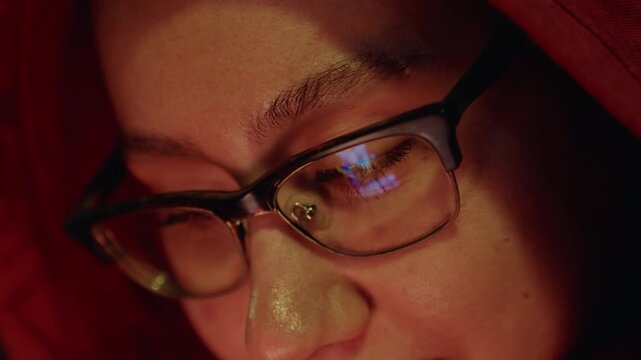 Asian woman glasses illuminated by screen, closeup portrait in red hoodie showing reflection on lenses, gentle smile, concentrated scrolling and tapping, study and coding vibes, messaging and social