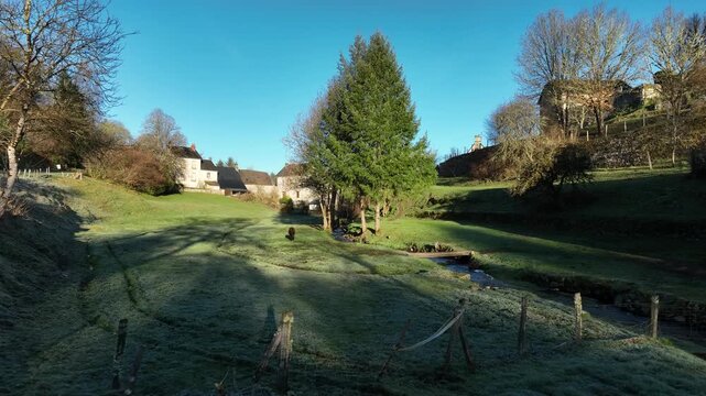 Frosty morning in rural Saint Angel Correze France with green meadows bare trees small stream and historic stone buildings under clear blue sky