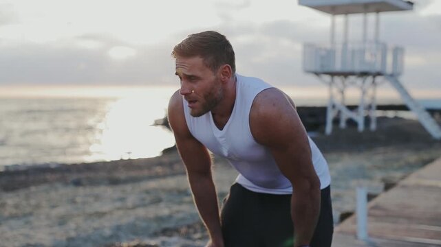 Young tired strong sporty athletic fit sportsman man wear white sports clothes taking respite breathing warm up train at sunrise sun dawn over sea beach outdoor on pier seaside in summer day morning