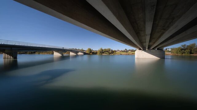 Concrete bridge stretching across a calm river, mirrored in smooth water beneath a bright blue sky with distant green trees along the serene banks