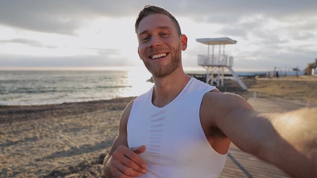 Young strong sporty athletic fit sportsman man wear sports clothes doing selfie shot pov on mobile cell phone warm up train at sunrise sun over sea beach outdoor on pier seaside in summer day morning