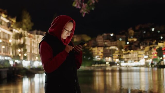 Woman in red hijab texting waterfront, soft phone glow illuminates face against bokeh city lights reflected on calm harbor water contemplative traveler checks messages under blossom branches,