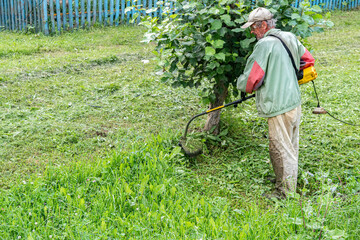 Obraz premium Senior man in cap cuts grass with an electric trimmer outside his house. He focuses on the task at hand