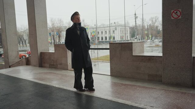 Young white creator on stone balcony studying rainy cityscape under portico wearing dark overcoat and beret slow contemplative turns textured paving distant buildings traffic cinematic moody