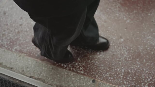 Closeup creator feet on terrazzo mat, dark trousers and polished leather shoes at entry threshold beside rubber grate, speckled floor texture and subtle motion, cinematic mood and intimate detail