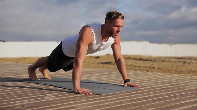 Full body young strong sporty athletic fit sportsman man he wear sports clothes do push-ups on yoga mat warm up train at sunrise sun dawn over sea beach outdoor on pier seaside in summer day morning