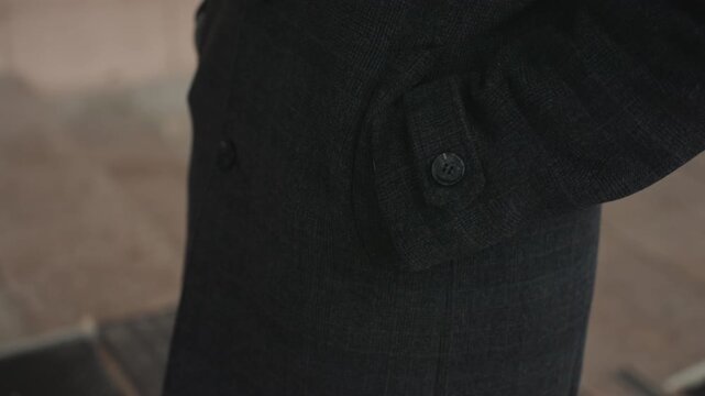 Closeup black coat pocket showing button and textured wool fabric, commuter adjusts sleeve on urban sidewalk muted tones, cinematic lighting emphasize tailoring, cuff and subtle gesture